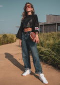A woman stands outdoors on a sandy path wearing a black wide crop top, blue jeans, white sneakers, and sunglasses, holding a football. A grassy field and a building are visible in the background.