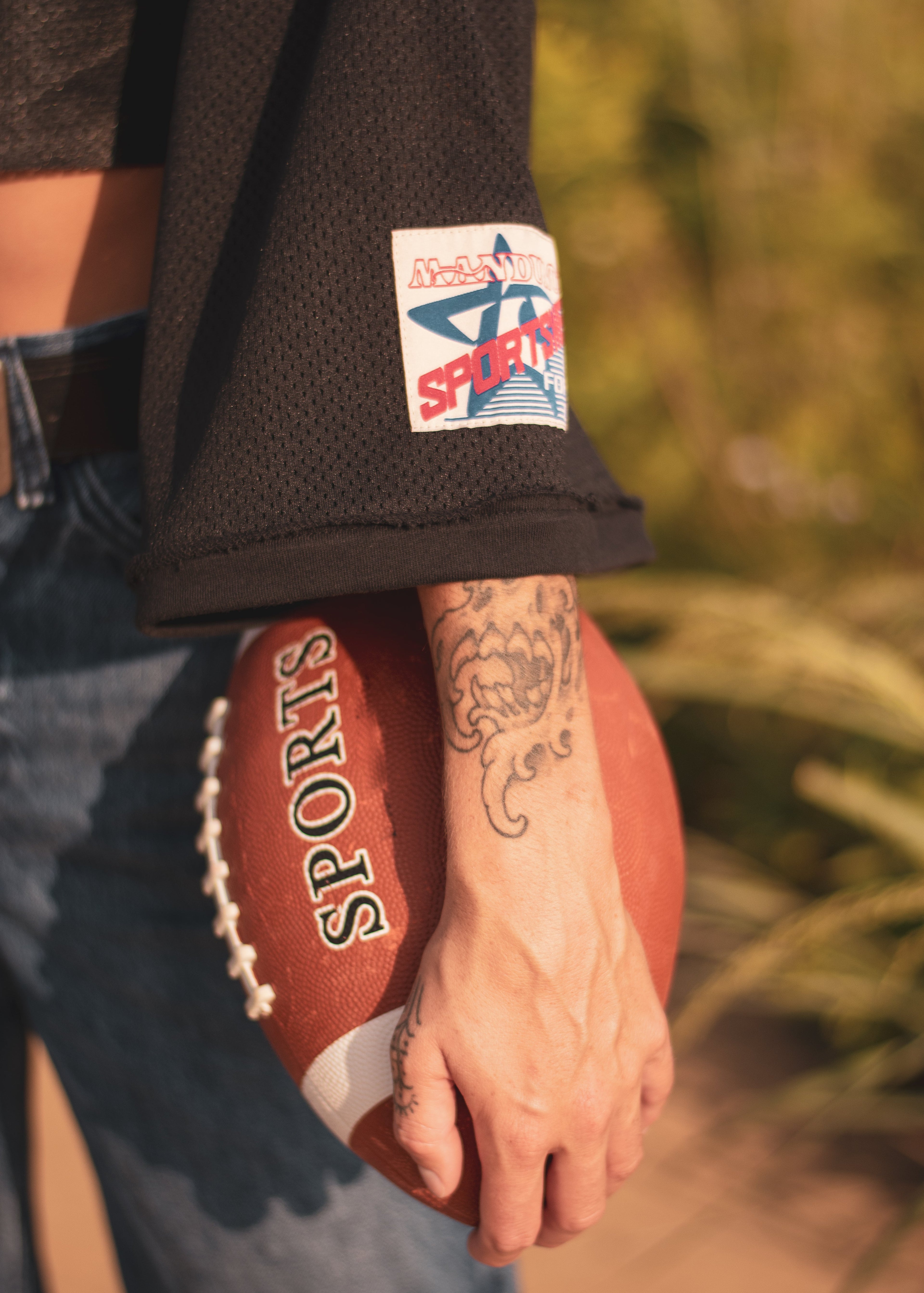 A close-up lifestyle shot of a person holding a brown leather football, wearing a black mesh sports jersey with a visible 'Manduno Sportswear' logo patch on the sleeve, set against a warm, blurred outdoor background.