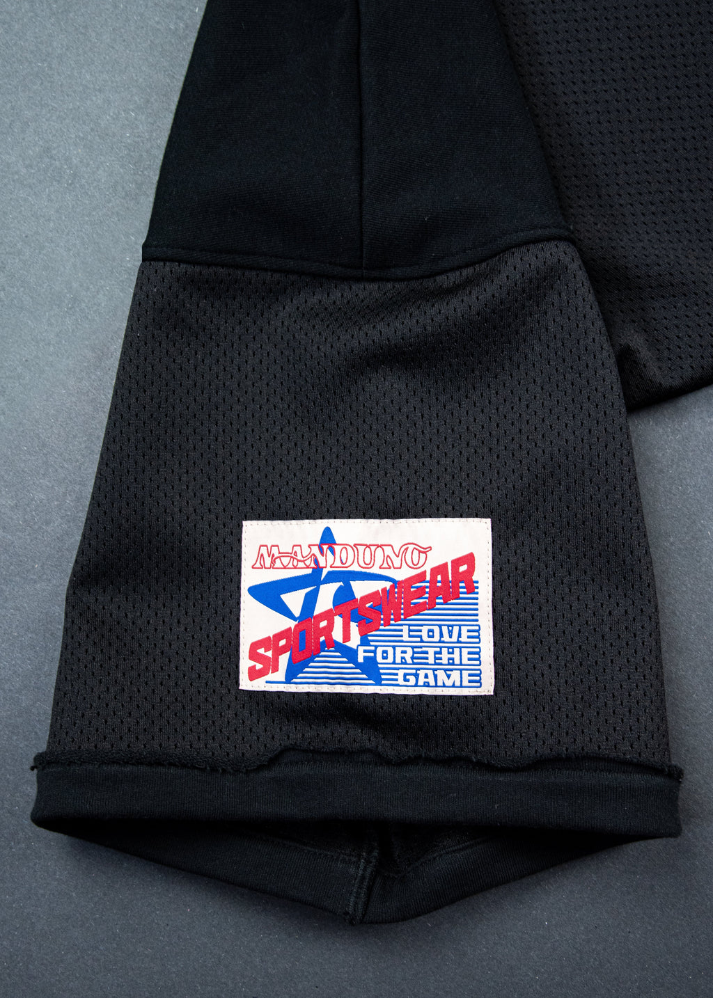 Close-up of a black mesh athletic jacket sleeve with a cuff, featuring a rectangular embroidered patch that reads 'Manduno Sportswear' in red, white, and blue.