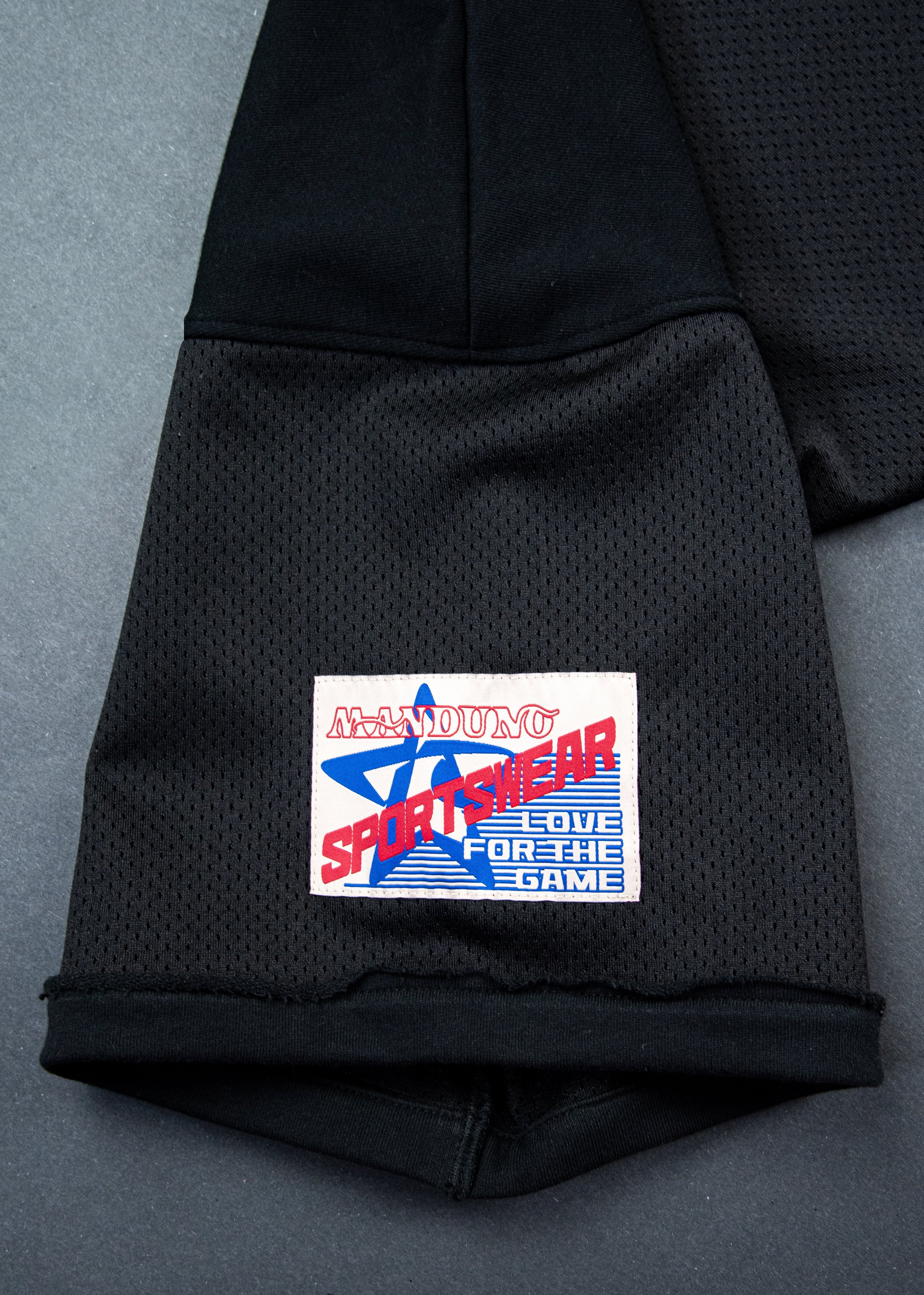 Close-up of a black mesh athletic jacket sleeve with a cuff, featuring a rectangular embroidered patch that reads 'Manduno Sportswear' in red, white, and blue.