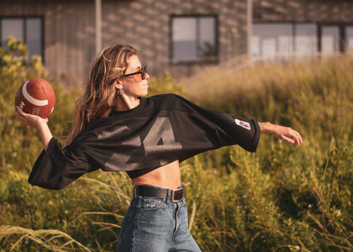 A woman outdoors in a grassy field wearing a black cropped long-sleeve shirt with a large black graphic on the front, blue jeans, and sunglasses, while holding a football.