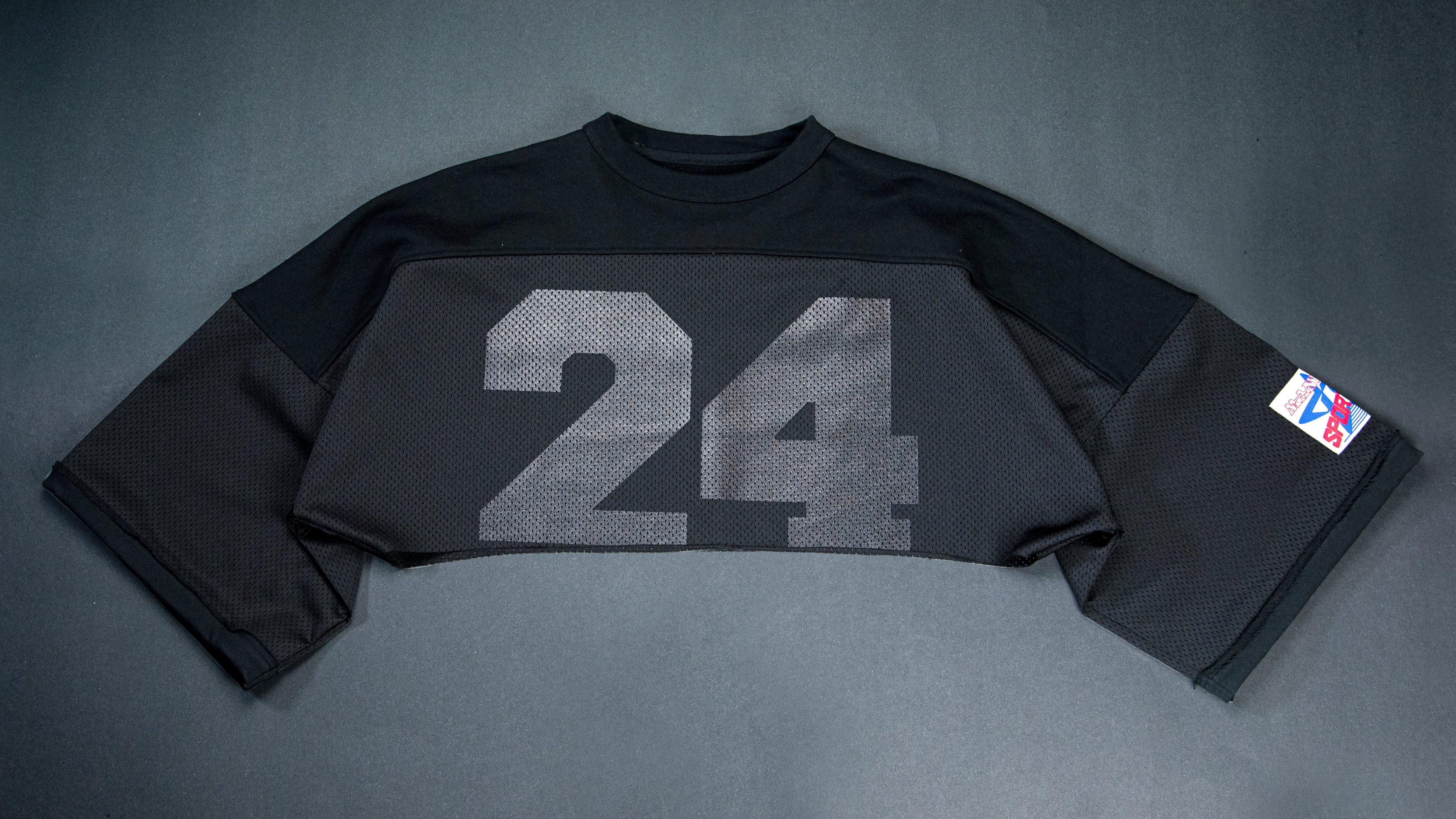 A black long-sleeve cropped jersey with a large gray '24' printed on the front and an NBA logo patch on the sleeve, laid flat on a dark gray background.