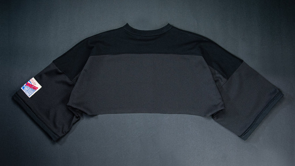 A black color-blocked long-sleeve crop top laid flat on a black background, featuring a small red, white, and blue patch on the left sleeve.