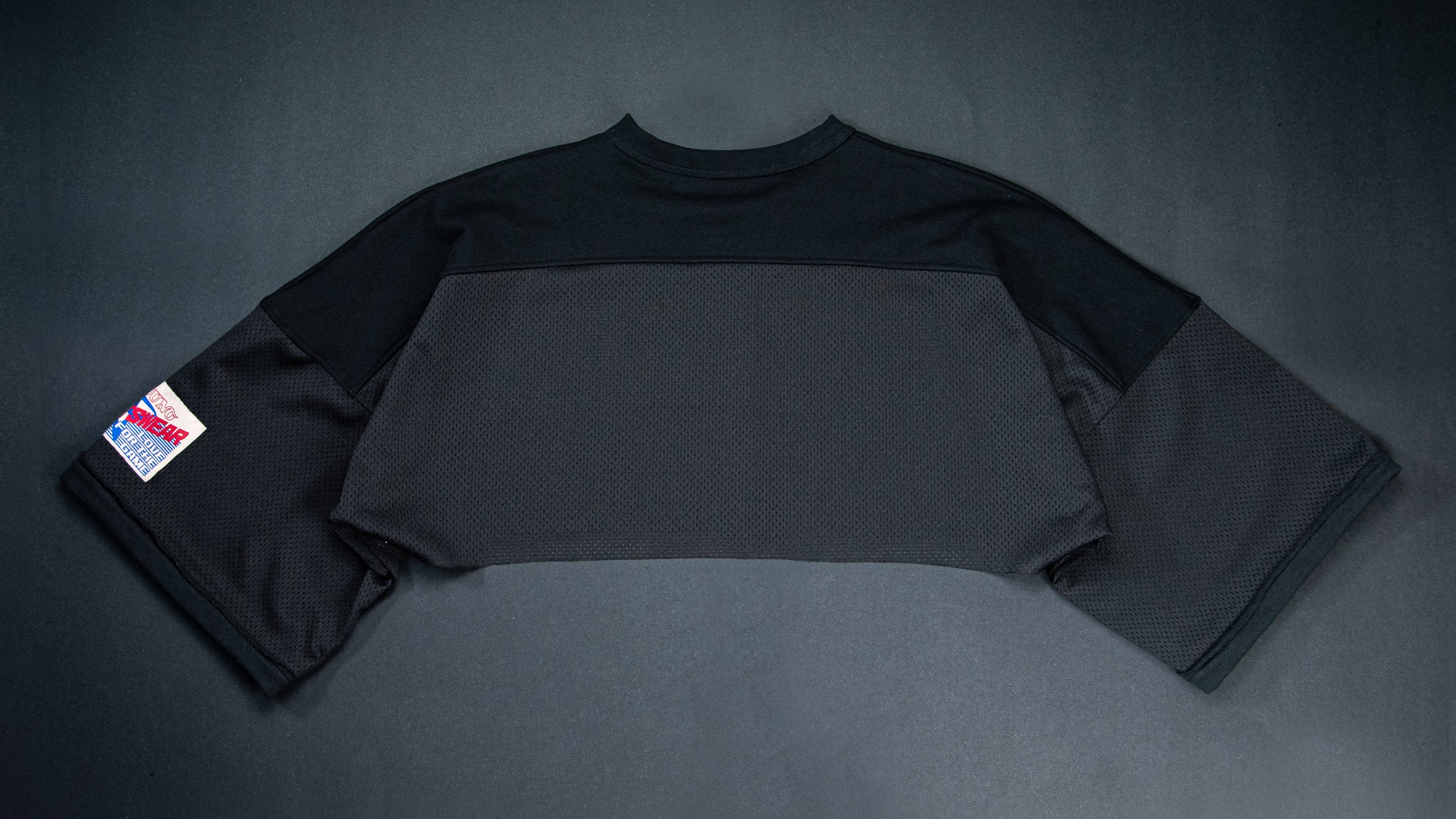 A black color-blocked long-sleeve crop top laid flat on a black background, featuring a small red, white, and blue patch on the left sleeve.