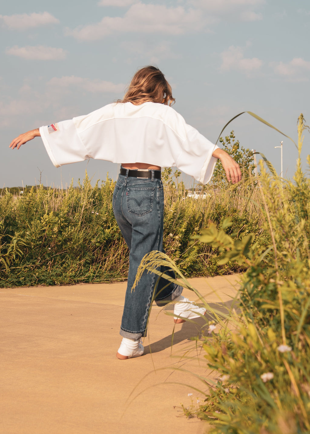 A woman wearing a white long-sleeve crop top and blue jeans is seen from behind, walking on a dirt path with her arms outstretched, surrounded by tall green grass under a blue sky.