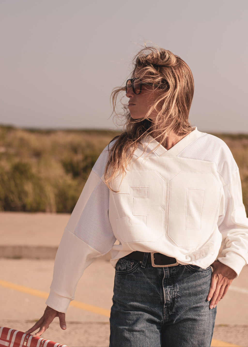 A woman wearing a white long-sleeve sweatshirt with textured applique numbers and mesh panels, paired with dark blue jeans, standing outdoors in a sunny, dry environment.