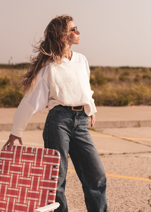 A woman wearing a white long-sleeve sweatshirt and dark blue jeans stands outdoors on a paved surface, holding a red and white patterned suitcase. She is wearing sunglasses, and the background shows a dry, grassy landscape under a hazy sky.