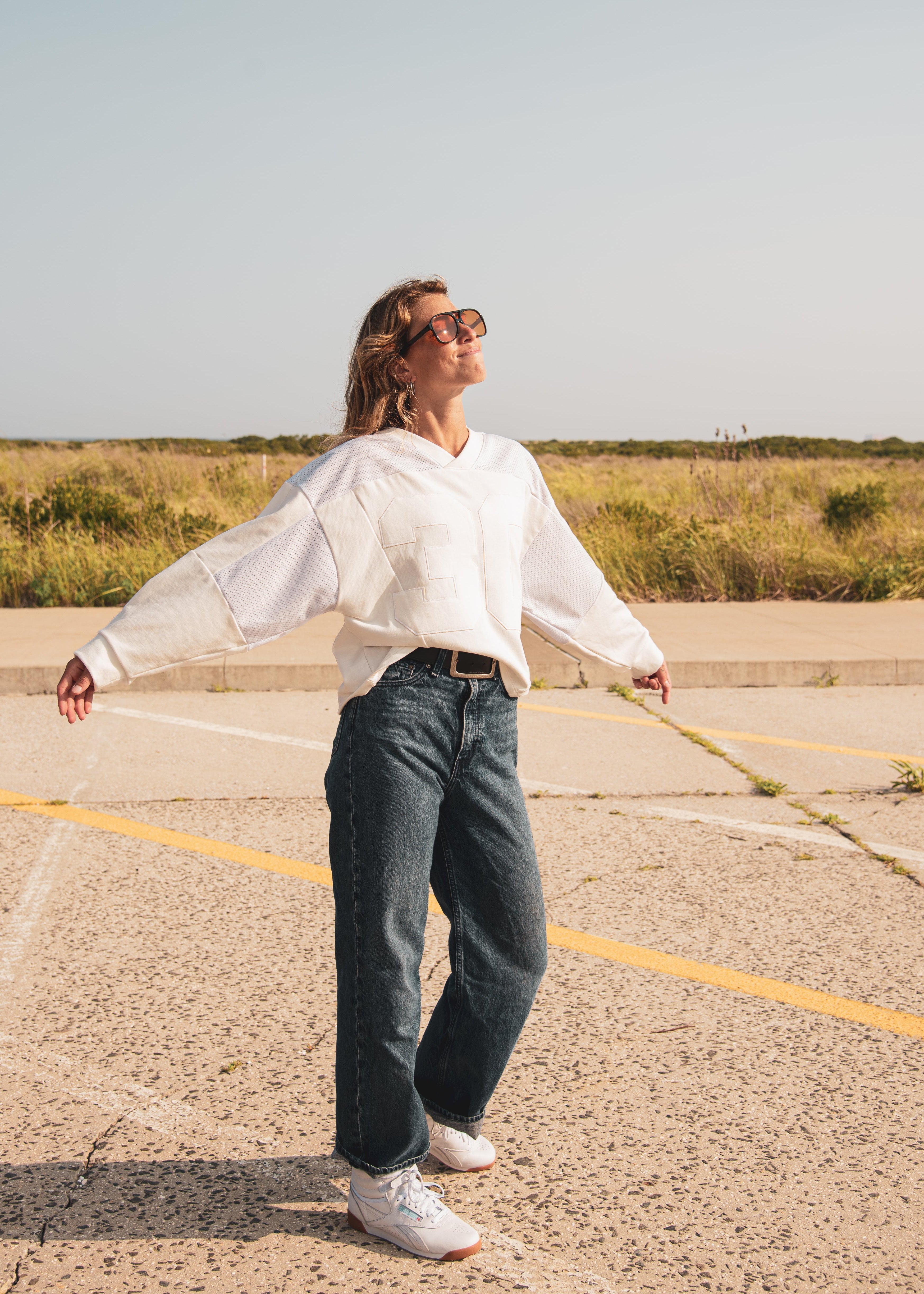 A woman wearing a white long-sleeve sweatshirt, dark blue wide-leg jeans, and white sneakers stands in an open paved area with grass in the background, looking upward with her arms outstretched.