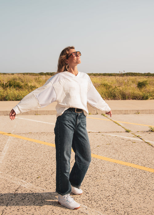 A woman wearing a white long-sleeve sweatshirt, dark blue wide-leg jeans, and white sneakers stands in an open paved area with grass in the background, looking upward with her arms outstretched.