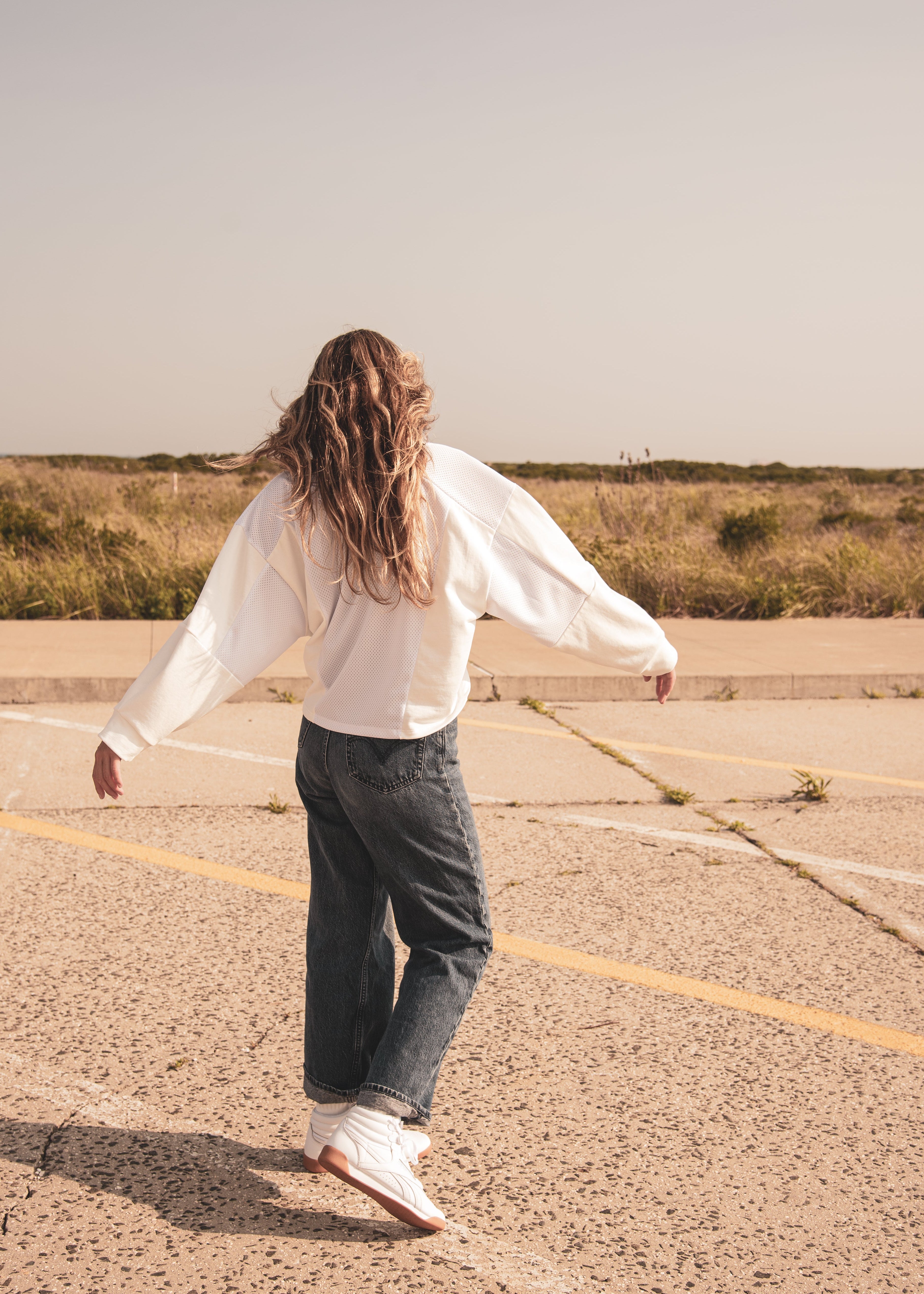 A person with long wavy brown hair seen from behind, wearing a white long-sleeve sweatshirt, dark gray jeans, and white sneakers, standing on a paved surface in an open outdoor area.
