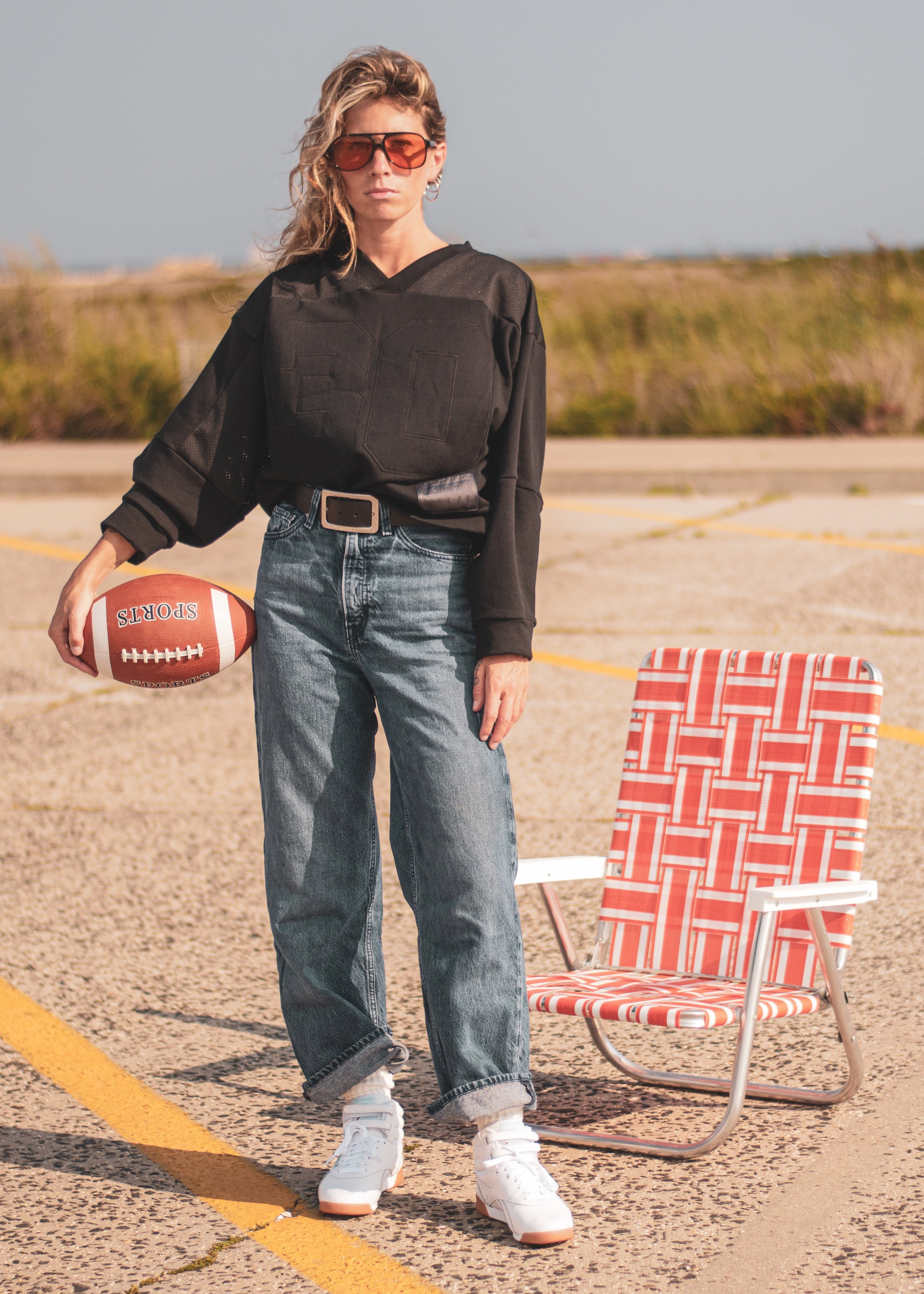 A woman stands on an outdoor paved surface wearing a black sweatshirt top, blue jeans, white sneakers, and sunglasses, holding a football next to a red-and-white patterned folding chair.