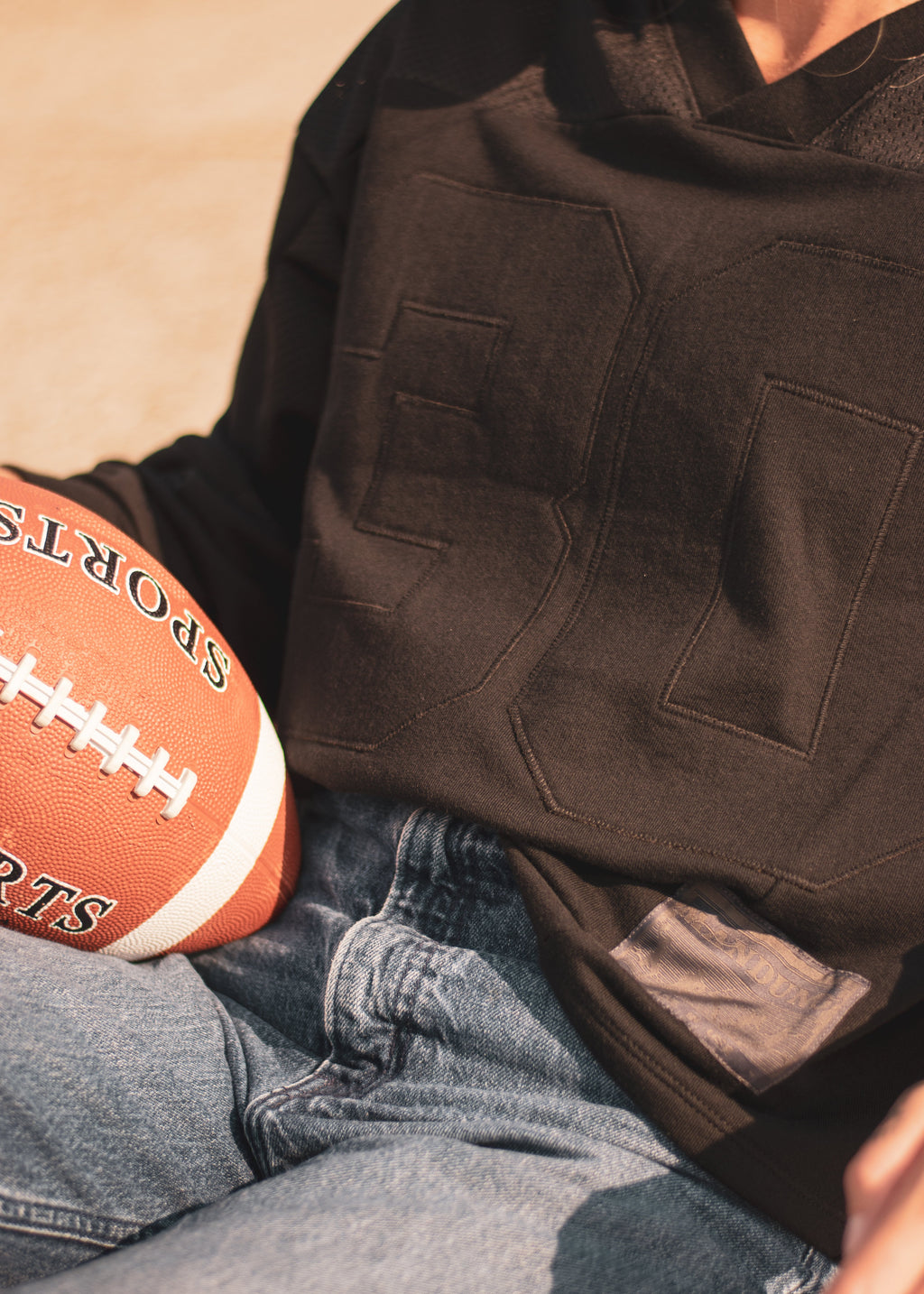 A close-up lifestyle shot of a person wearing a black long-sleeve sweatshirt with a large applique numbers paired with blue jeans, holding a brown and white football.
