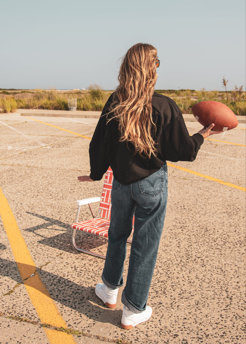 A woman stands in a sunny outdoor parking lot holding a football, viewed from behind. She is wearing a black long-sleeve top, blue jeans, and white sneakers, with a red-and-white striped folding chair beside her.
