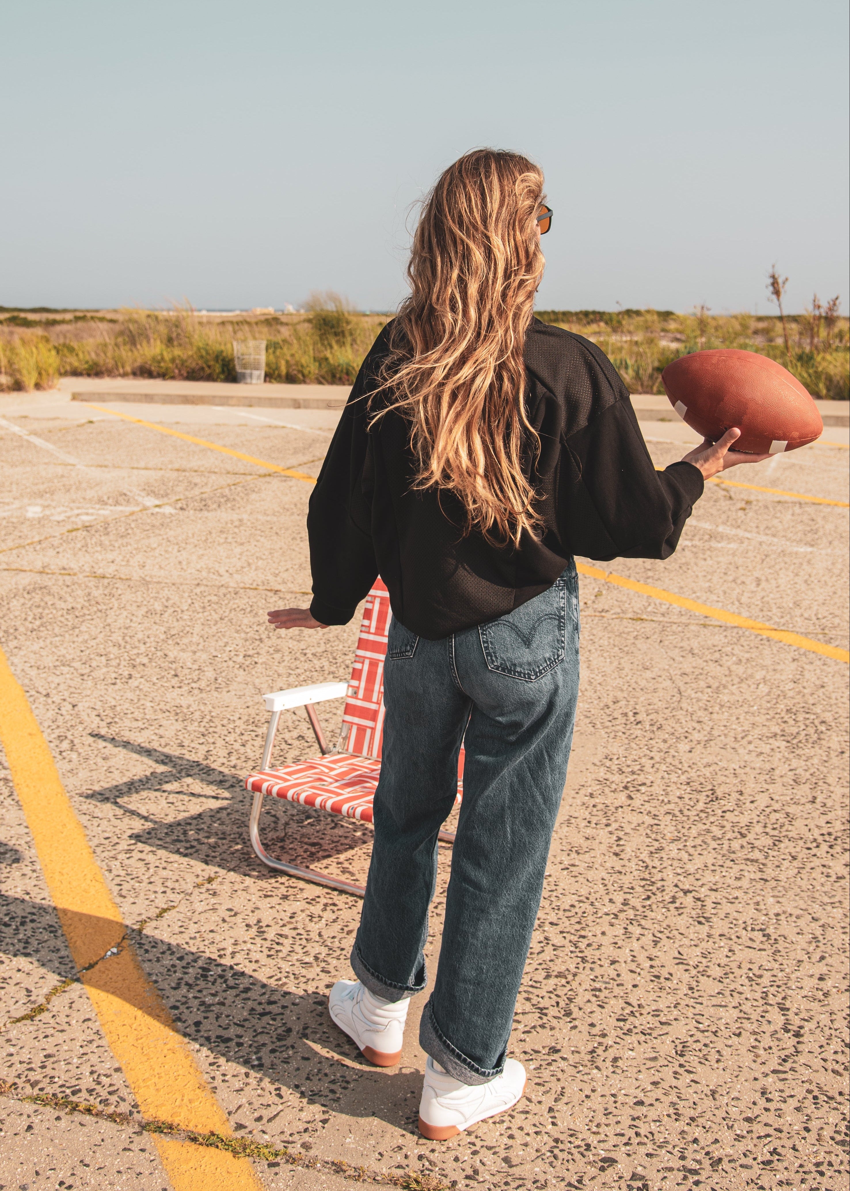 A woman stands in a sunny outdoor parking lot holding a football, viewed from behind. She is wearing a black long-sleeve top, blue jeans, and white sneakers, with a red-and-white striped folding chair beside her.