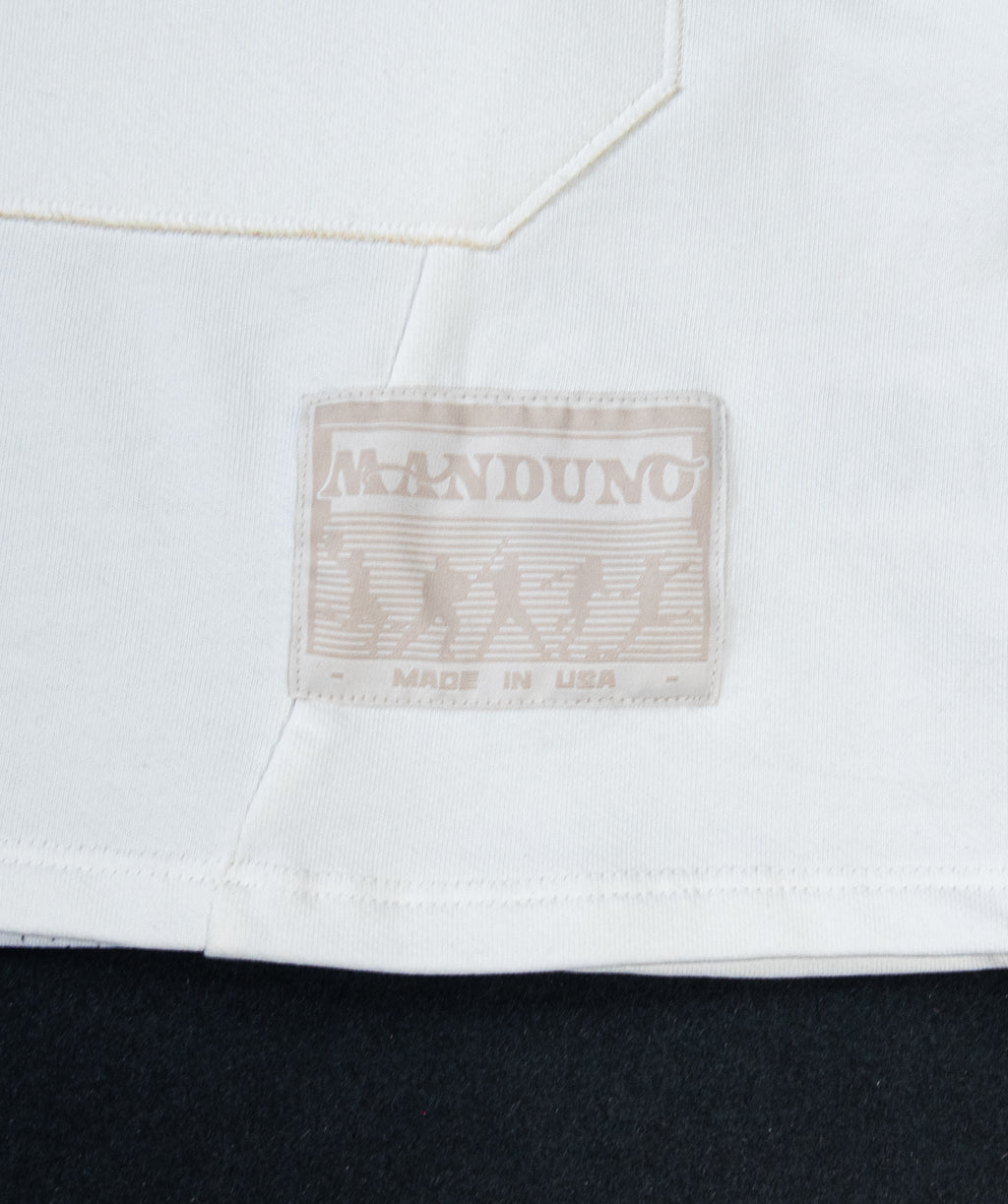 Close-up of a white garment with a sewn-on brand patch reading MANDUNO and MADE IN USA, shown against a dark background.