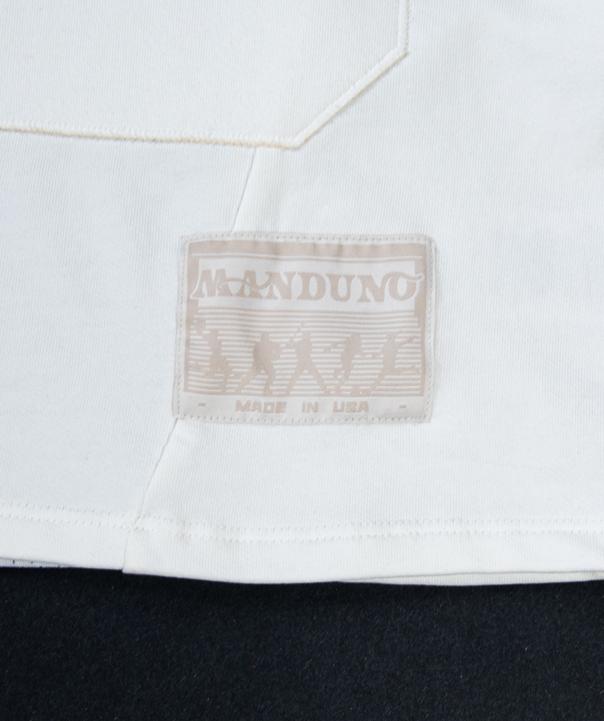 Close-up of a white garment with a sewn-on brand patch reading MANDUNO and MADE IN USA, shown against a dark background.