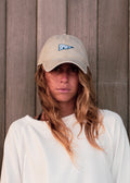 A woman with long light brown hair wears a beige baseball cap with a blue and blu 'SPORTS' flag logo and a white sweatshirt, standing against a vertical wooden plank wall.