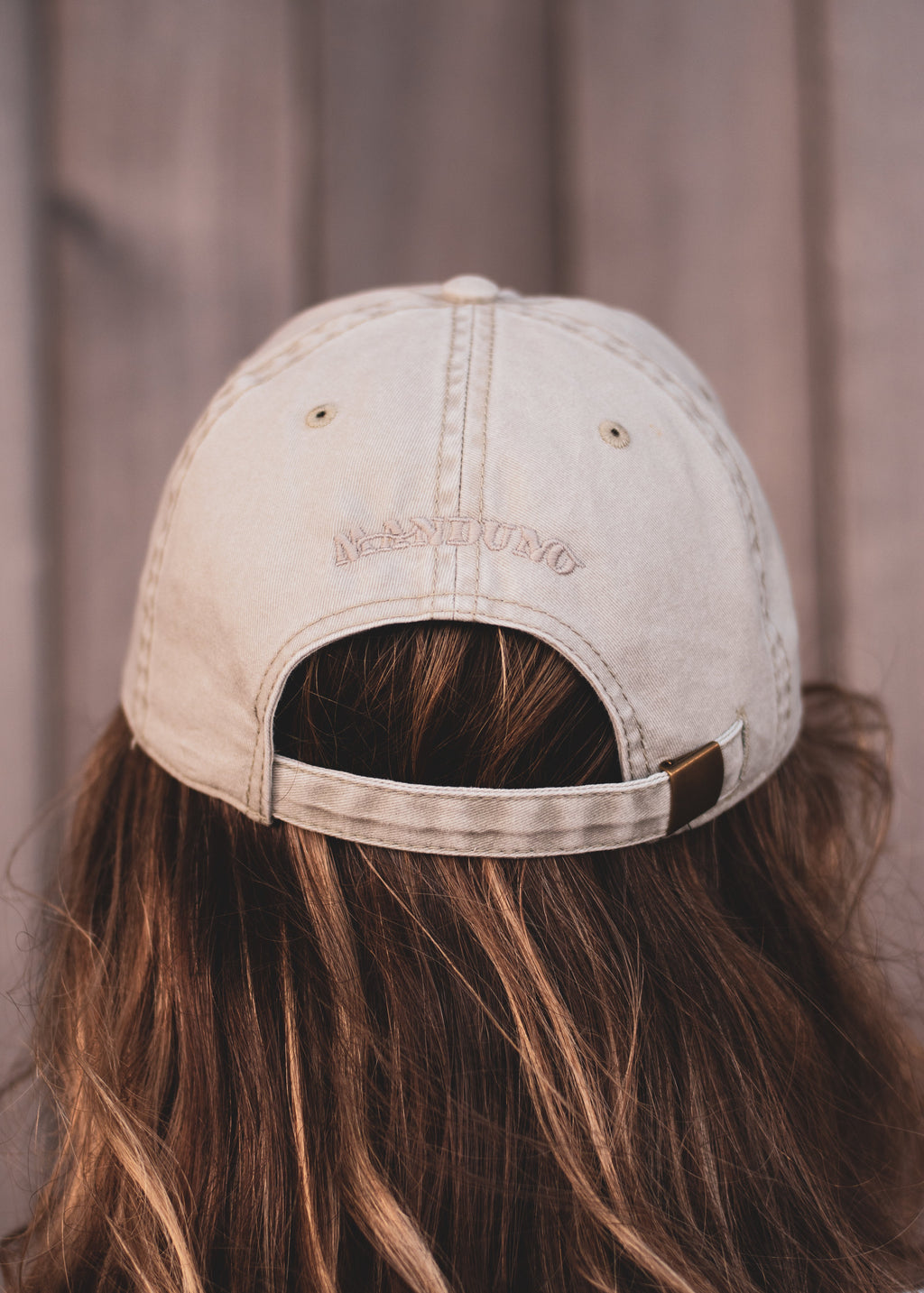 A person viewed from behind wearing a light beige washed baseball cap with 'MANDUNO' embroidered on the back, set against a soft-focus brown wooden background.