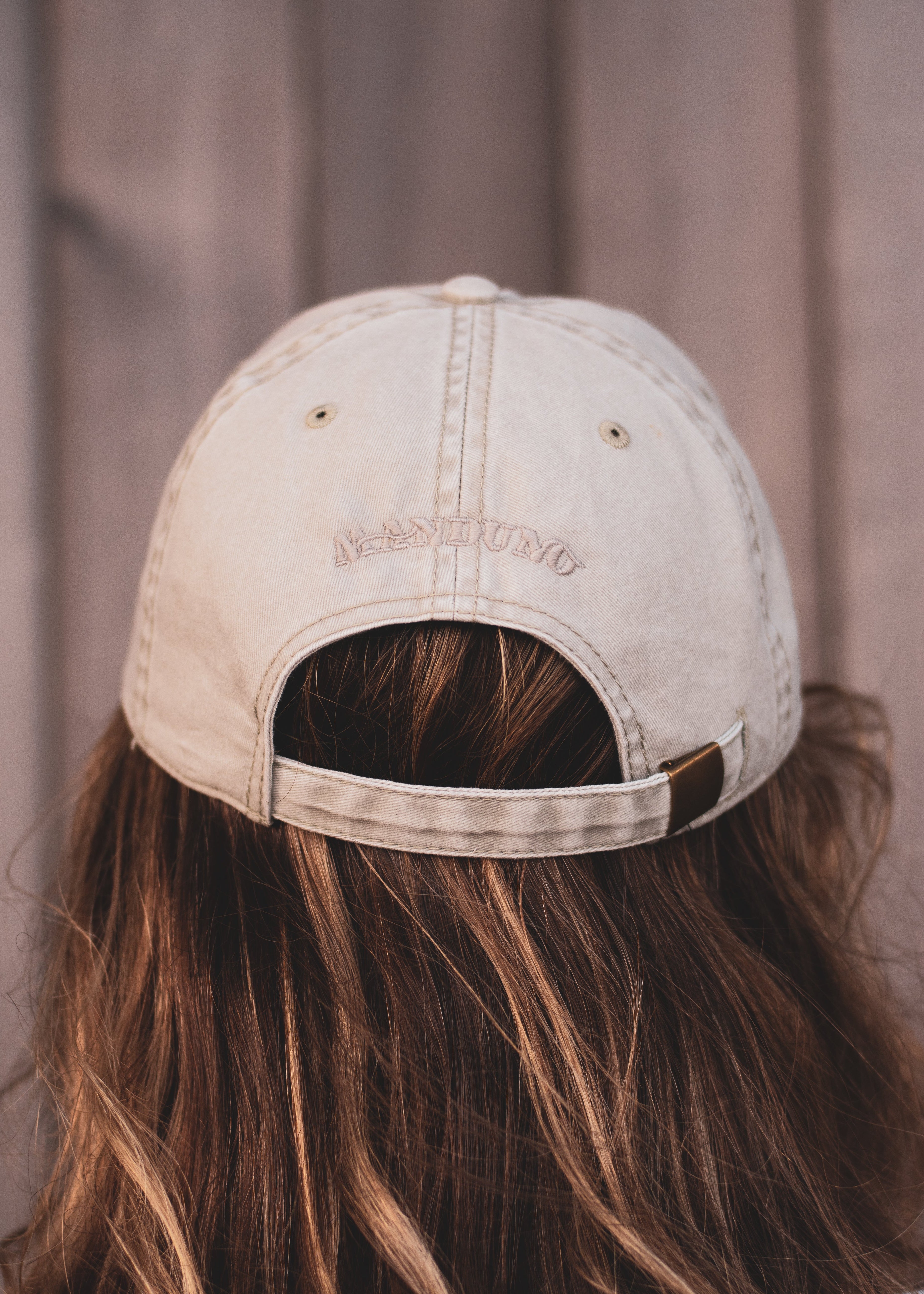 A person viewed from behind wearing a light beige washed baseball cap with 'MANDUNO' embroidered on the back, set against a soft-focus brown wooden background.