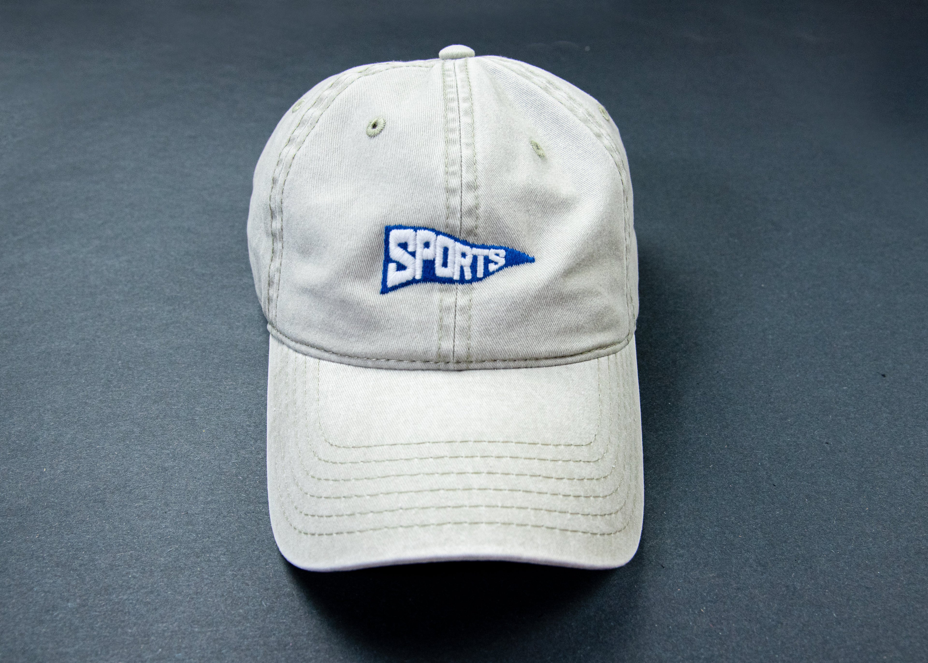 A light beige baseball cap with a blue and white embroidered 'SPORTS' pennant logo on the front, displayed on a dark gray background.