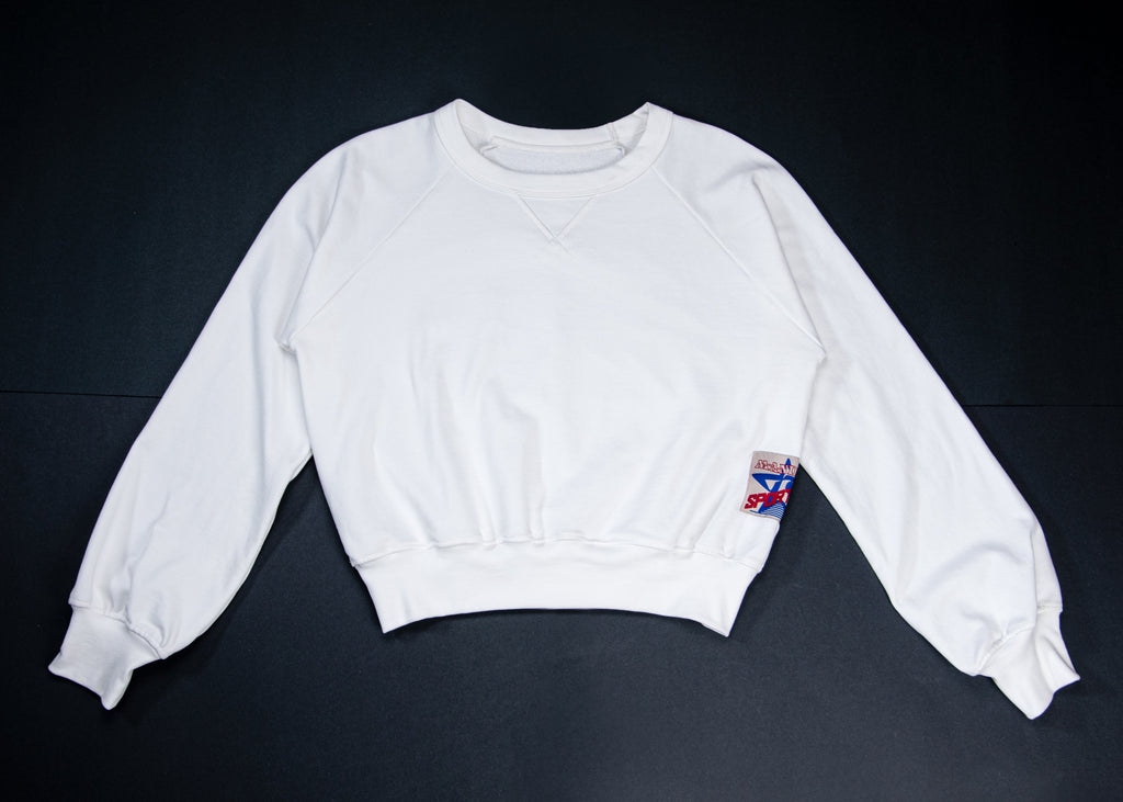 A white long-sleeve cropped sweatshirt laid flat on a dark background, featuring a small colorful patch on the left sleeve.