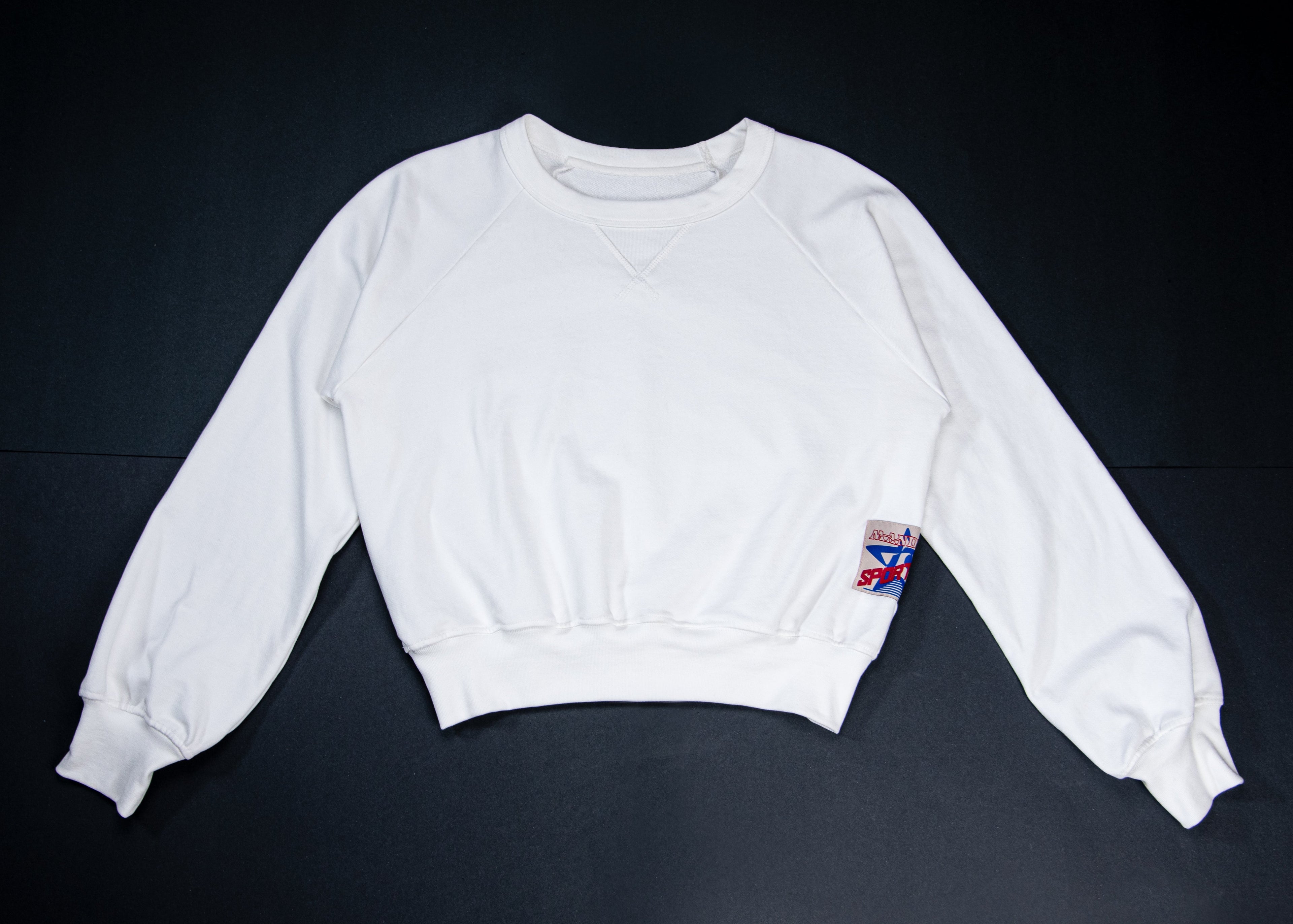 A white long-sleeve cropped sweatshirt laid flat on a dark background, featuring a small colorful patch on the left sleeve.