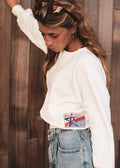 A woman wearing a white long-sleeve crewneck sweatshirt with a vintage-style 'Sportswear' patch on the bottom hem, paired with light-wash blue jeans, posed against a rustic wooden plank wall.