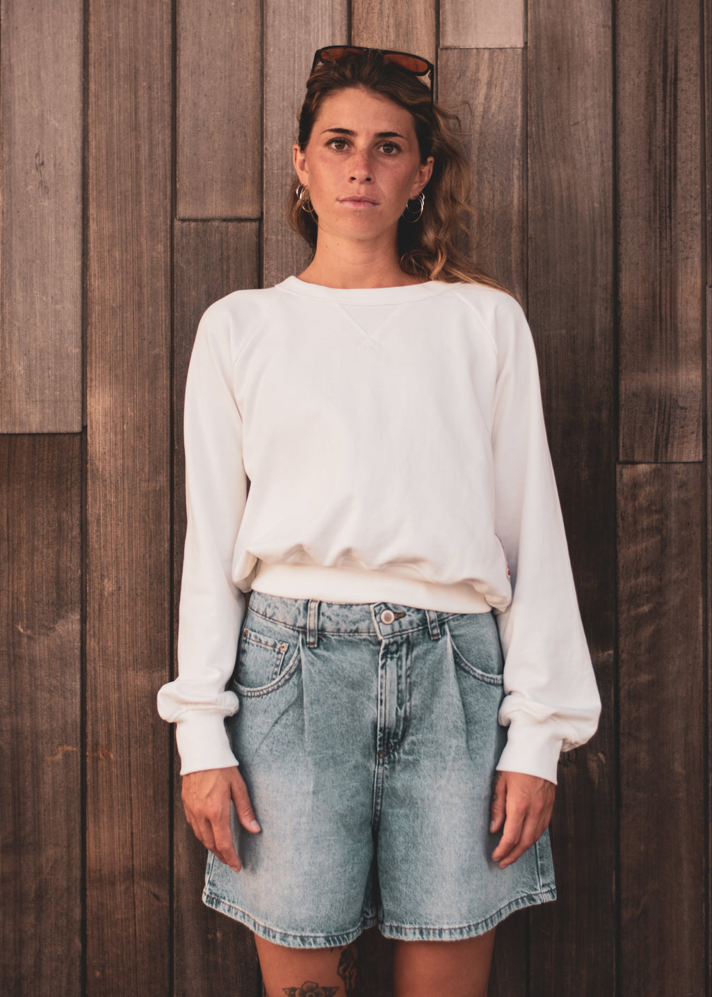 A woman stands against a dark wood plank wall wearing a white long-sleeve crewneck sweatshirt and light-wash denim shorts.