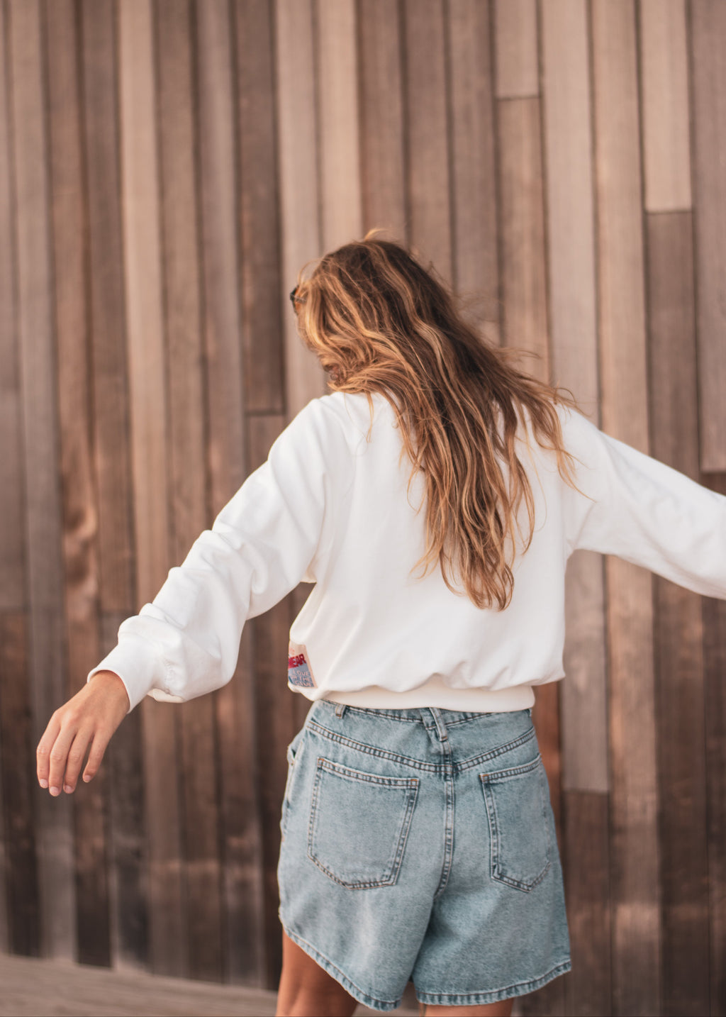 A woman with long light brown hair is shown from behind, wearing a white long-sleeve sweatshirt and light-wash denim shorts, standing in front of a vertical wooden plank wall.
