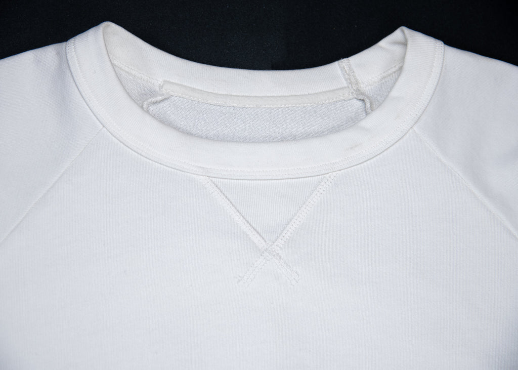 Close-up of a white crewneck sweatshirt showing the ribbed collar, front neck gusset, and shoulder seams against a black background.
