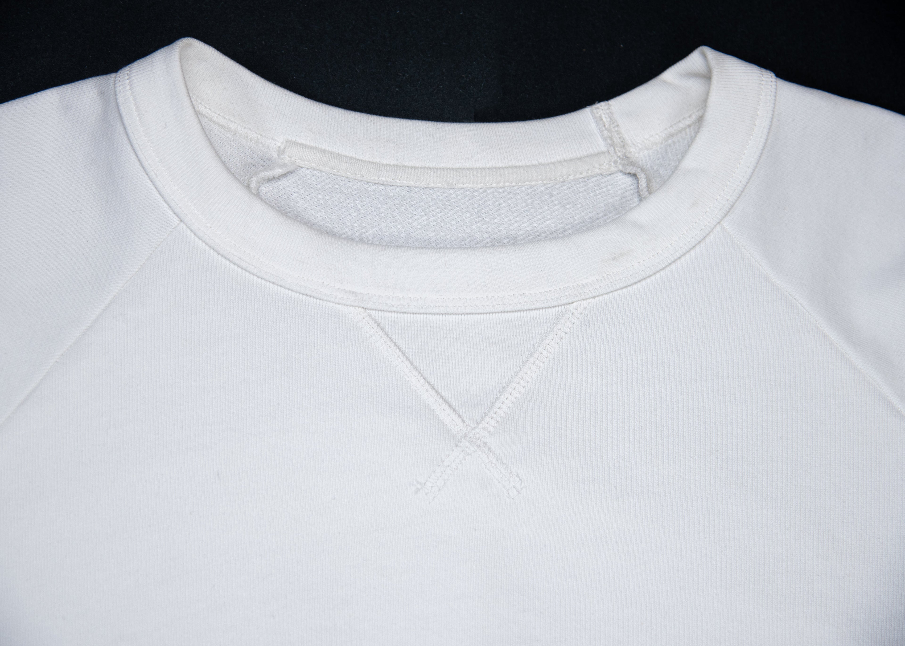 Close-up of a white crewneck sweatshirt showing the ribbed collar, front neck gusset, and shoulder seams against a black background.
