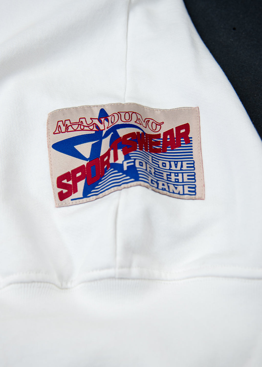 Close-up of a white garment featuring a rectangular woven patch on the side. The patch has a beige background with a blue star, red text reading 'MADUNO SPORTSWEAR', and blue striped text reading 'FOR LOVE FOR THE GAME'.