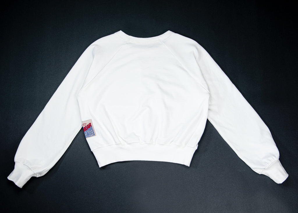 A white long-sleeve sweatshirt laid flat against a dark background, showing the back view with a small red and blue tag near the left sleeve.