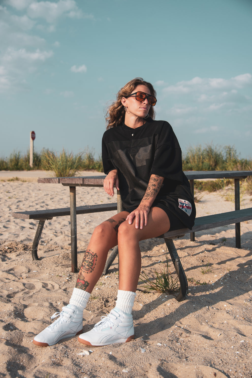 A woman sits on a wooden picnic table at a sandy beach, wearing a black oversized t-shirt, black shorts, white socks, and white sneakers.