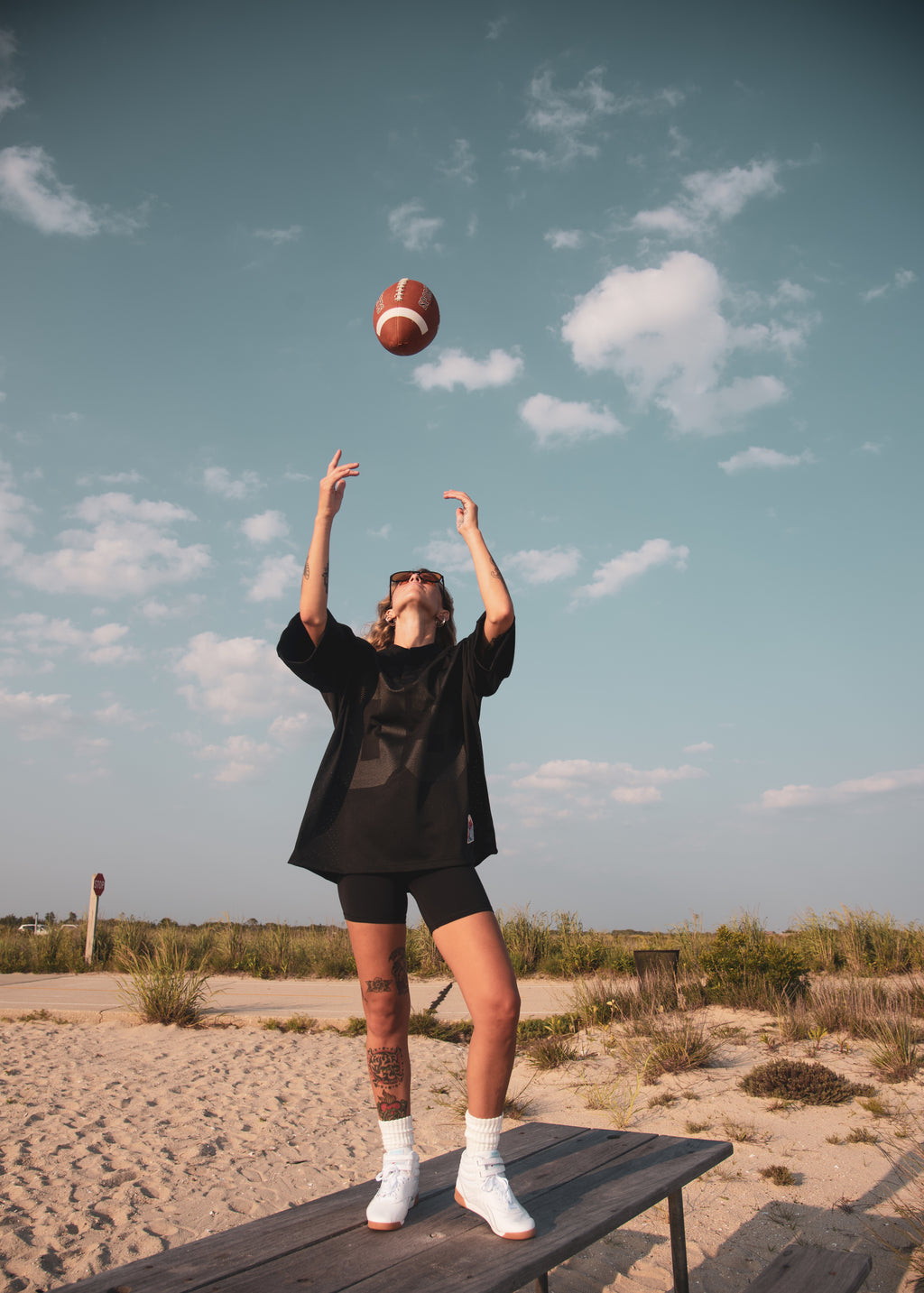 A person standing on a wooden bench outdoors in a sandy, grassy area under a blue sky, tossing a football overhead. They are wearing an oversized black t-shirt, black shorts, white socks, and white sneakers.