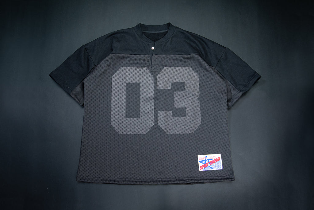 A black on black short-sleeve henley-style shirt with a large gray '03' printed on the front, laid flat against a dark background. A small red, white, and blue patch is visible near the lower right hem.