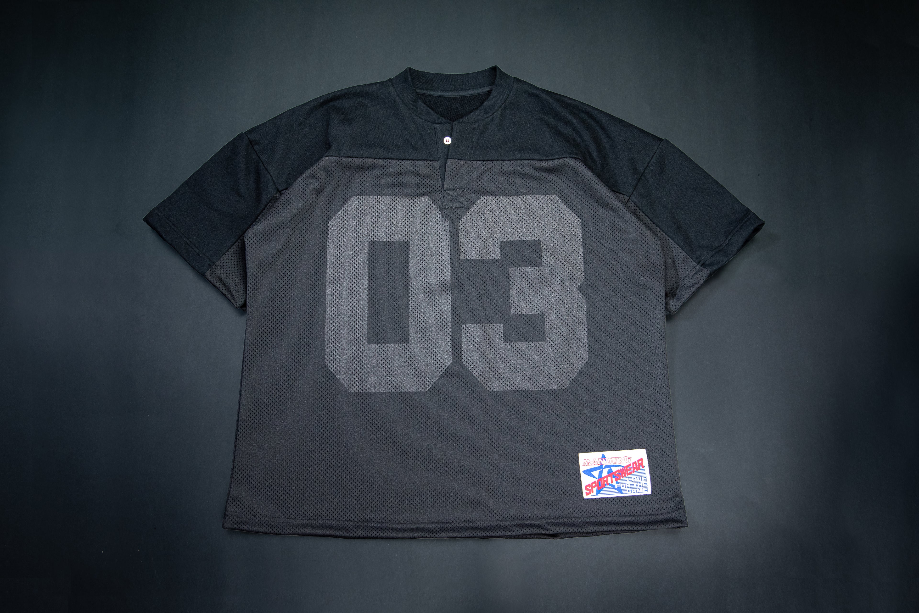 A black on black short-sleeve henley-style shirt with a large gray '03' printed on the front, laid flat against a dark background. A small red, white, and blue patch is visible near the lower right hem.