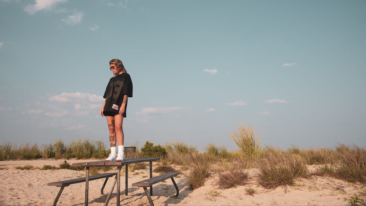 A person stands on a picnic table in an outdoor sandy dune setting under a large blue sky, wearing a black oversized T-shirt, black shorts, and white sneakers.
