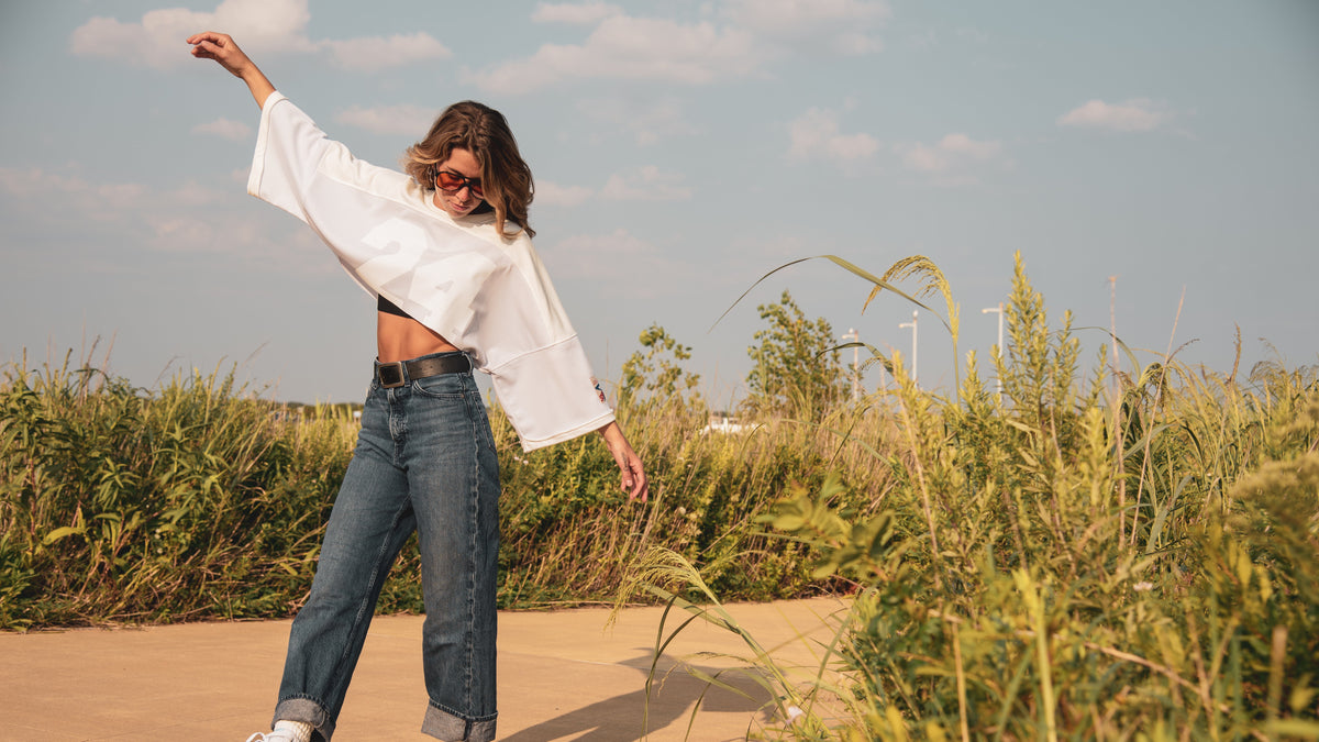 A woman outdoors in a field wearing a white cropped long-sleeve shirt, blue jeans, and white sneakers, captured in a sunny lifestyle shot.