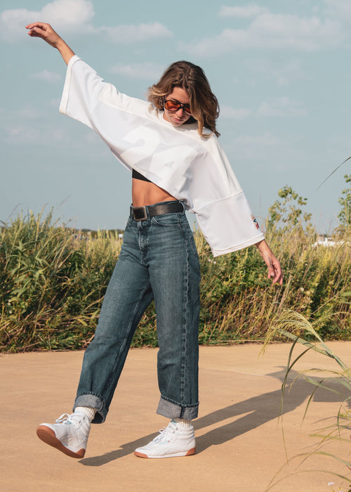 A woman posing outdoors in a white cropped long-sleeve shirt, blue jeans, and white sneakers on a sandy path surrounded by tall grass under a blue sky.