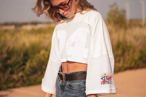 A woman wearing a white cropped mesh long-sleeve shirt with a large white number 24 on the front and a small Manduno Sportswear patch on the sleeve, paired with blue jeans and sunglasses, outdoors.