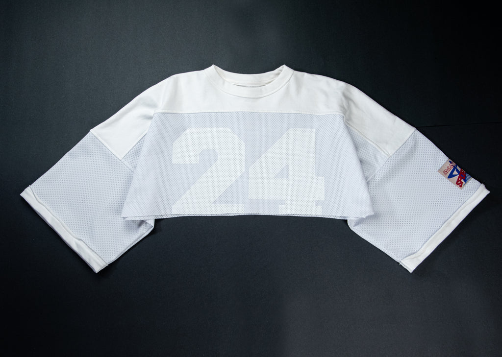A white long-sleeve football-style jersey laid flat on a dark background, featuring a large white '24' on the front and a small multicolor patch on the sleeve.