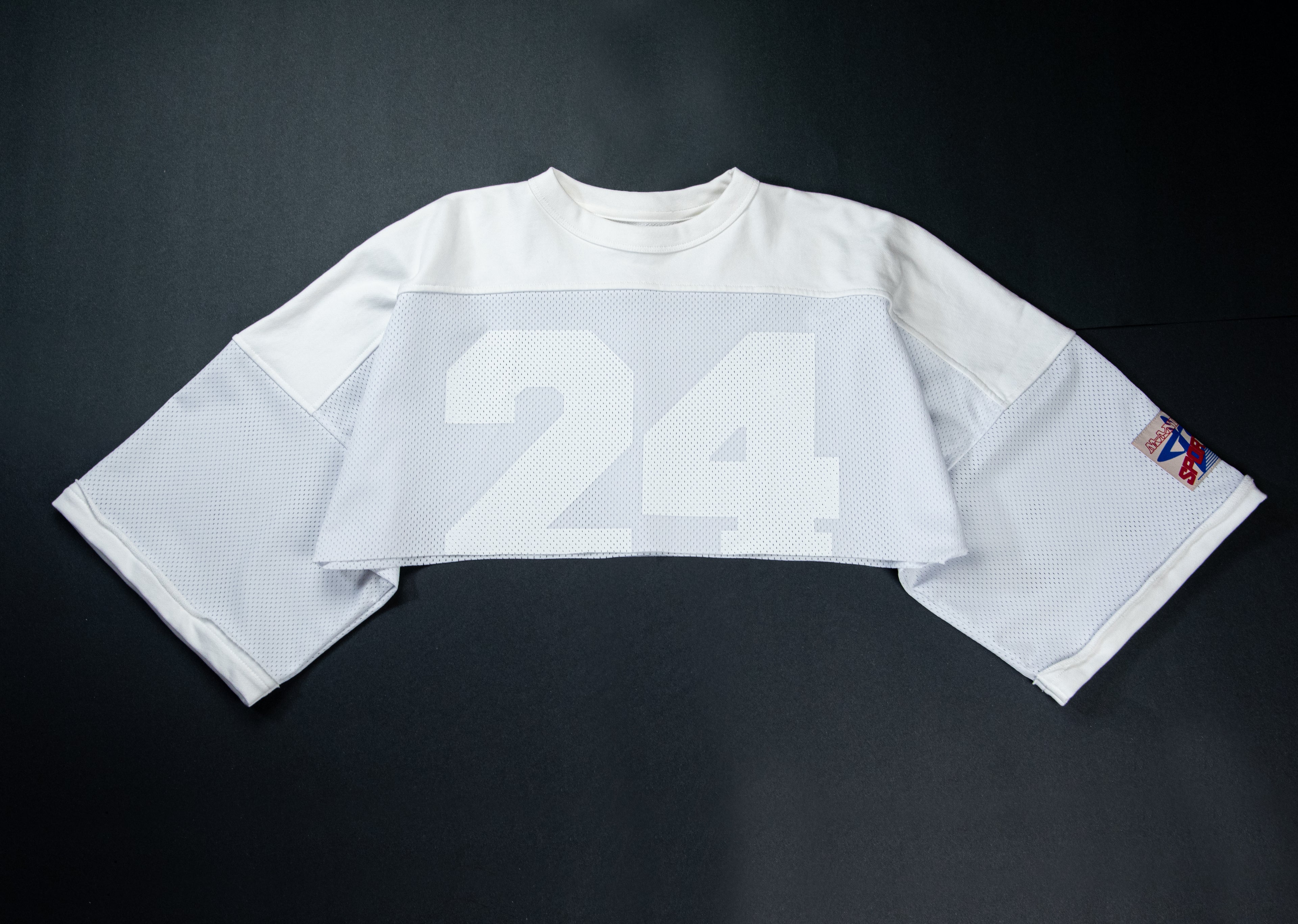 A white long-sleeve football-style jersey laid flat on a dark background, featuring a large white '24' on the front and a small multicolor patch on the sleeve.