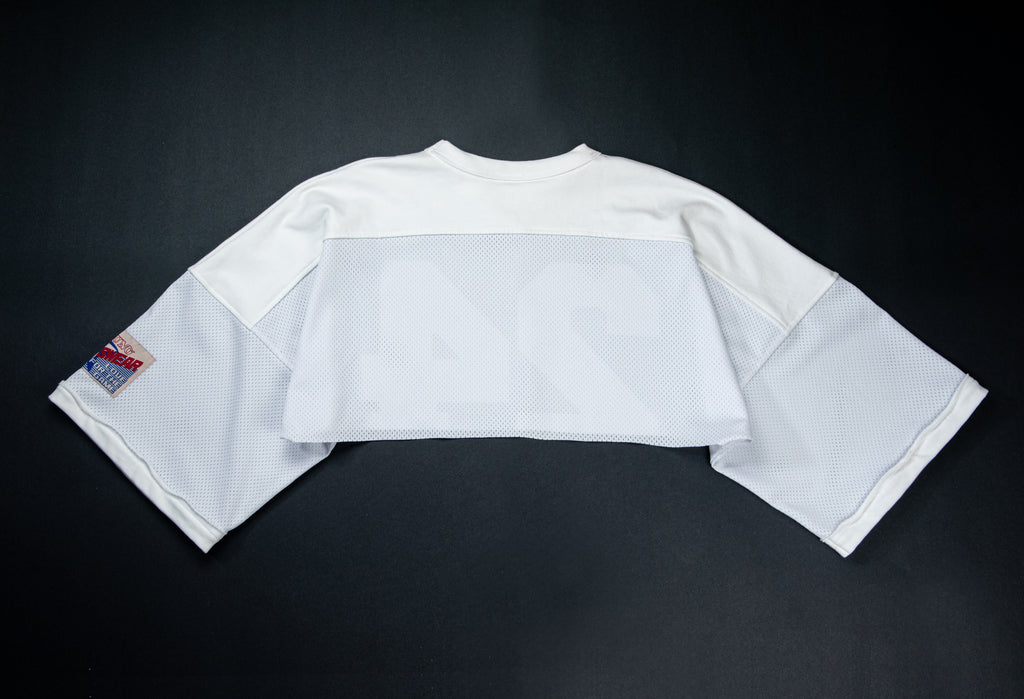 A white cropped football jersey with 3/4 sleeves, laid flat on a dark background. The jersey features a solid white upper chest and shoulders, a semi-sheer white mesh band across the chest, and a small red and blue patch on the right sleeve.
