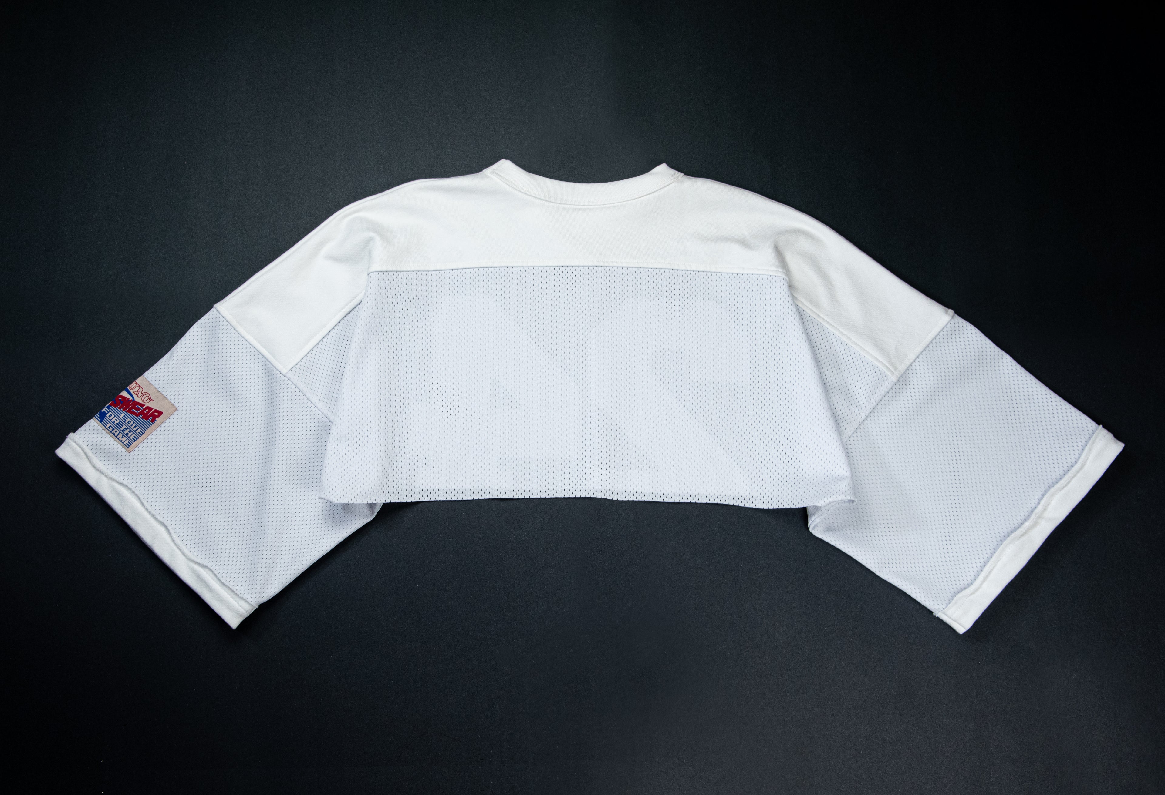 A white cropped football jersey with 3/4 sleeves, laid flat on a dark background. The jersey features a solid white upper chest and shoulders, a semi-sheer white mesh band across the chest, and a small red and blue patch on the right sleeve.