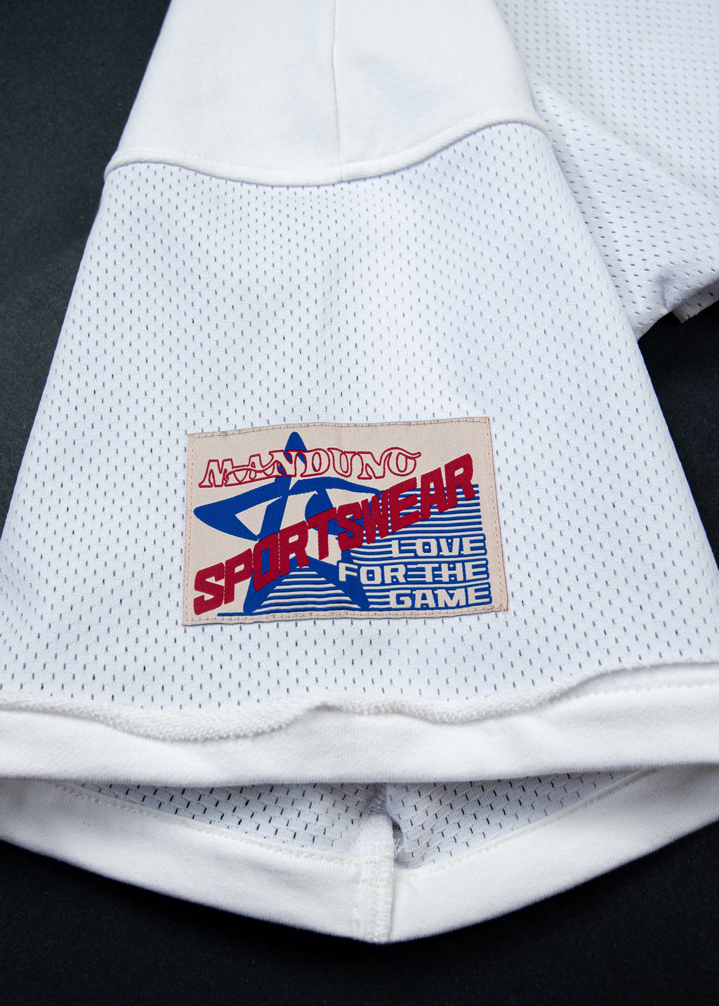 Close-up of a white mesh athletic jersey with a rectangular embroidered patch reading 'MANDUNO SPORTWEAR' and 'LOVE FOR THE GAME' on a black background.
